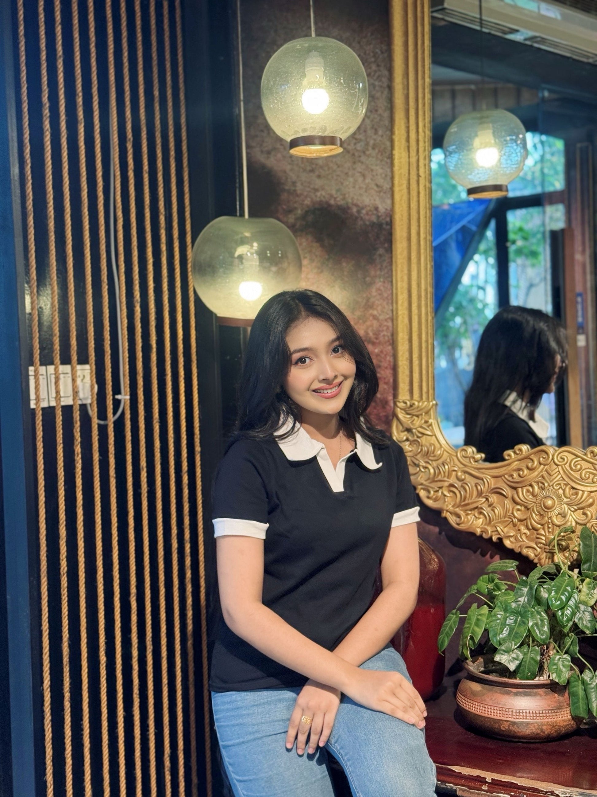 Woman sitting in a cafe with decorative mirrors and lights in the background in wearing our Black & White contrast Polo T-Shirt