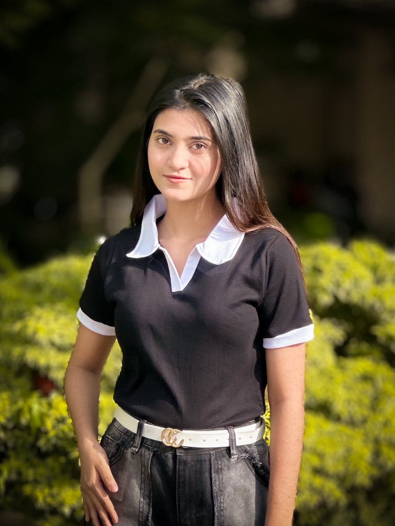 Woman wearing a black polo shirt with white collar and trim, standing outdoors with greenery in the background.