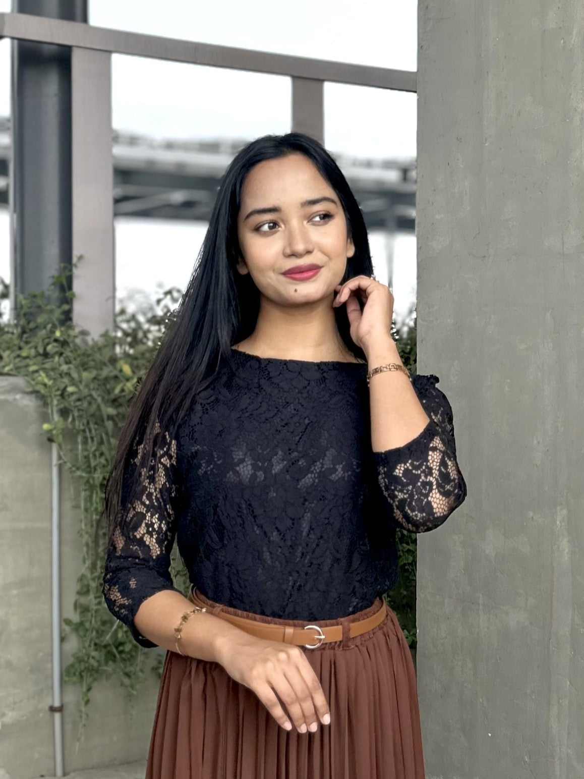 Woman wearing a black lace top and brown skirt standing against a neutral background