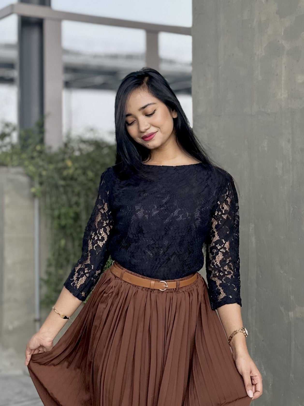Woman wearing a black lace top and brown skirt standing against a concrete wall.