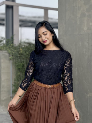 Woman wearing a black lace top and brown skirt standing against a concrete wall.