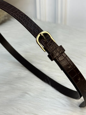 A close-up of a dark brown leather belt with a crocodile-embossed texture and a polished gold-tone buckle, resting on a white faux fur surface. rendy Buzz BD.