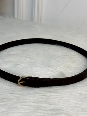 A close-up of a dark brown leather belt with a crocodile-embossed texture and a polished gold-tone buckle, resting on a white faux fur surface. rendy Buzz BD.