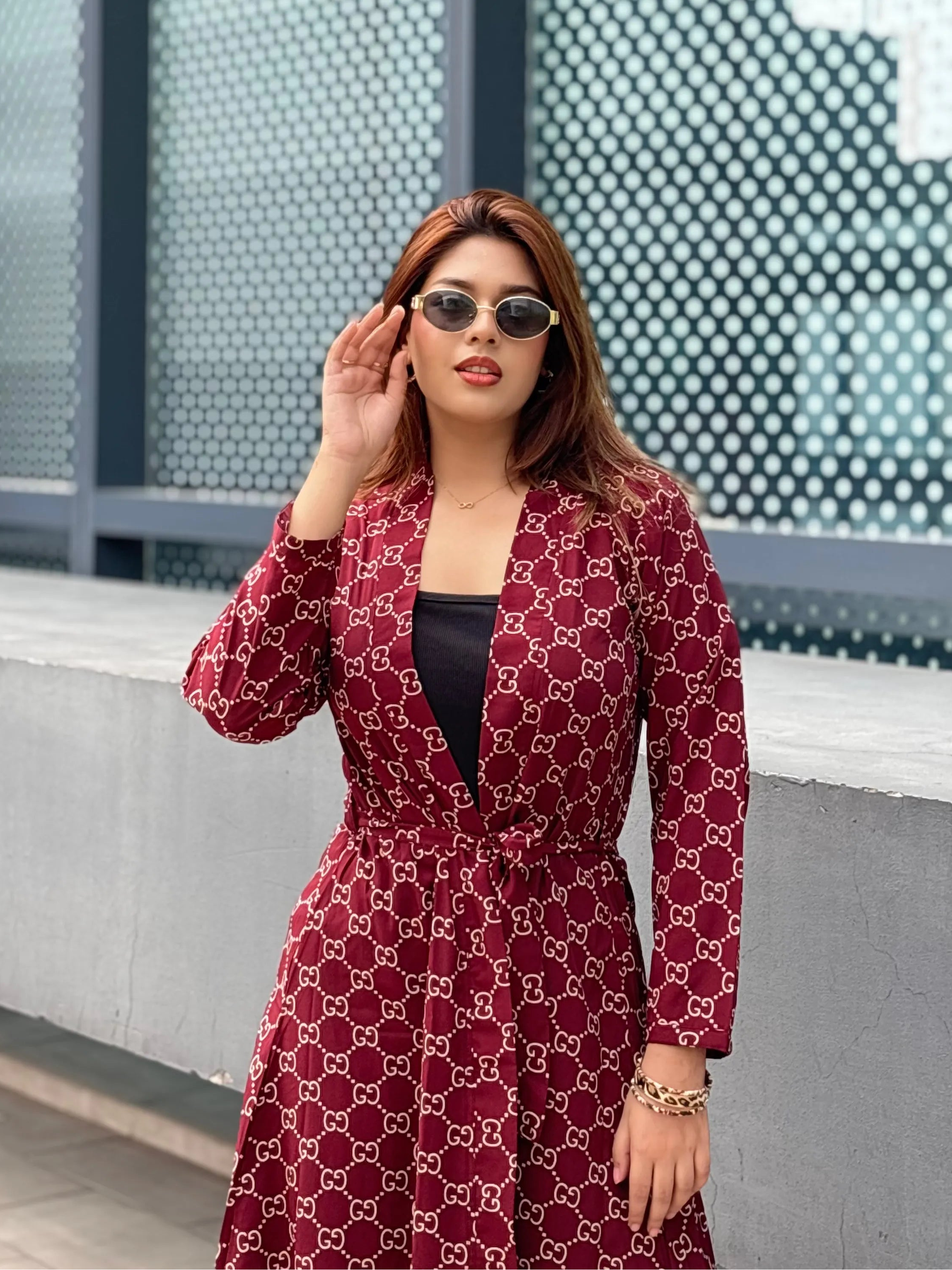 Woman wearing a red patterned Shrug and sunglasses, standing against a modern building facade.