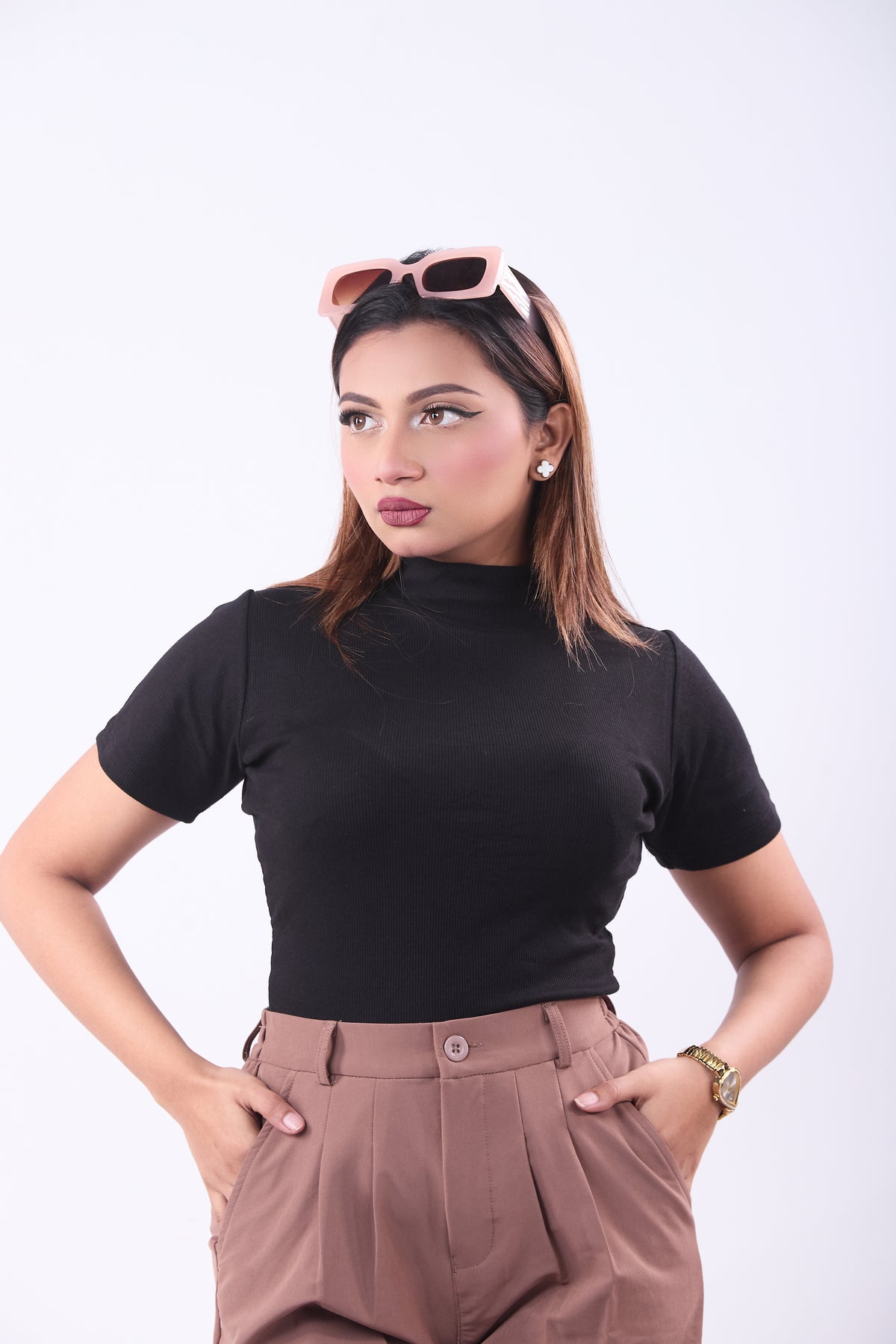 Woman wearing a black turtleneck and brown pants with sunglasses on her head against a white background