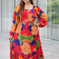 Woman wearing a colorful floral dress standing indoors with large windows.