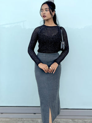 Woman wearing a black transparent top and gray pencil skirt against a light blue wall.