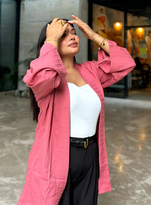 A woman posing in a pink crinkled-texture open-front Kimono over a white top and black pants, standing in a modern indoor/outdoor corridor.