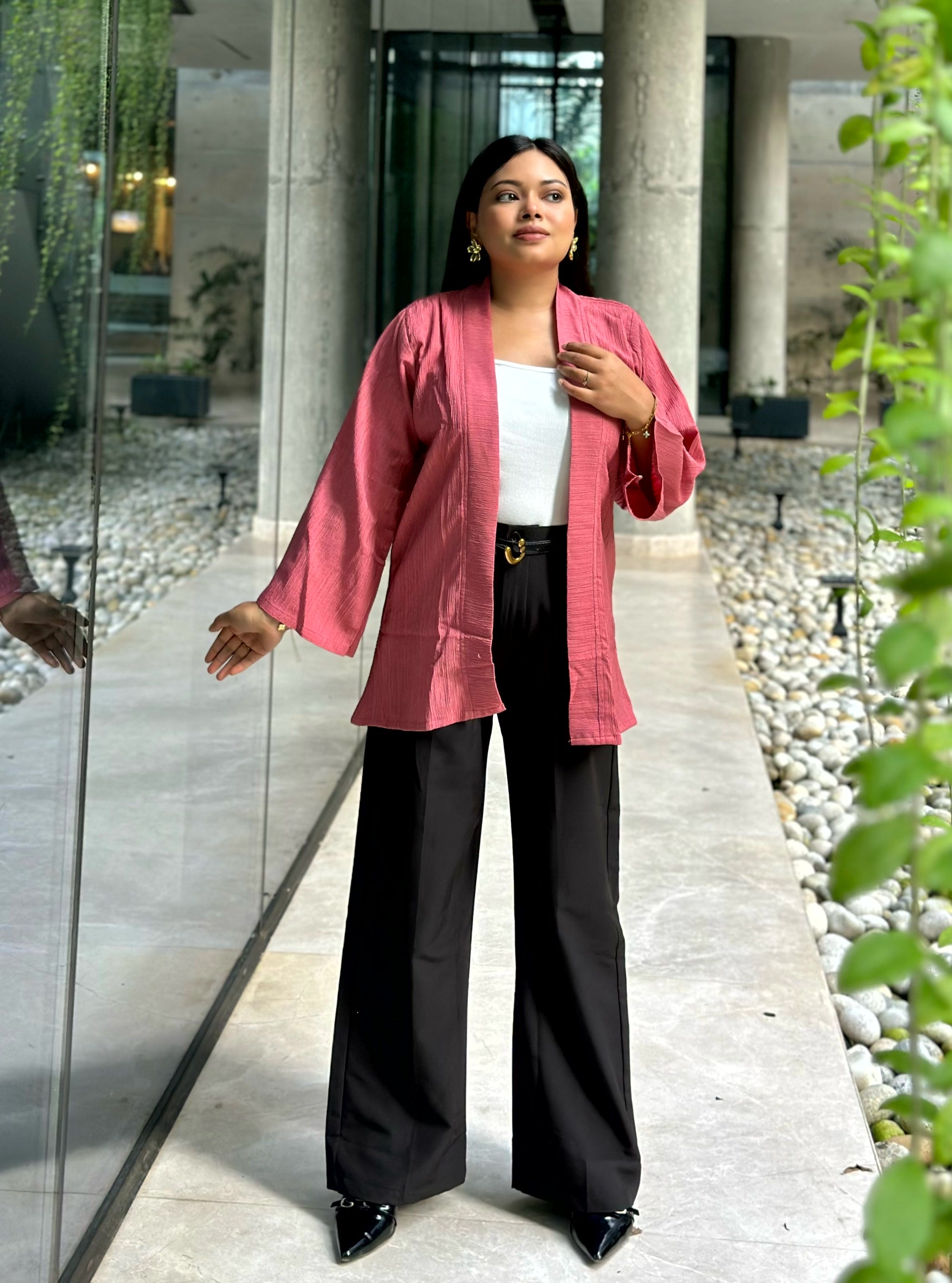 A woman stands in a modern outdoor walkway wearing a pink open-front Kimono over a white top and black wide-leg pants.