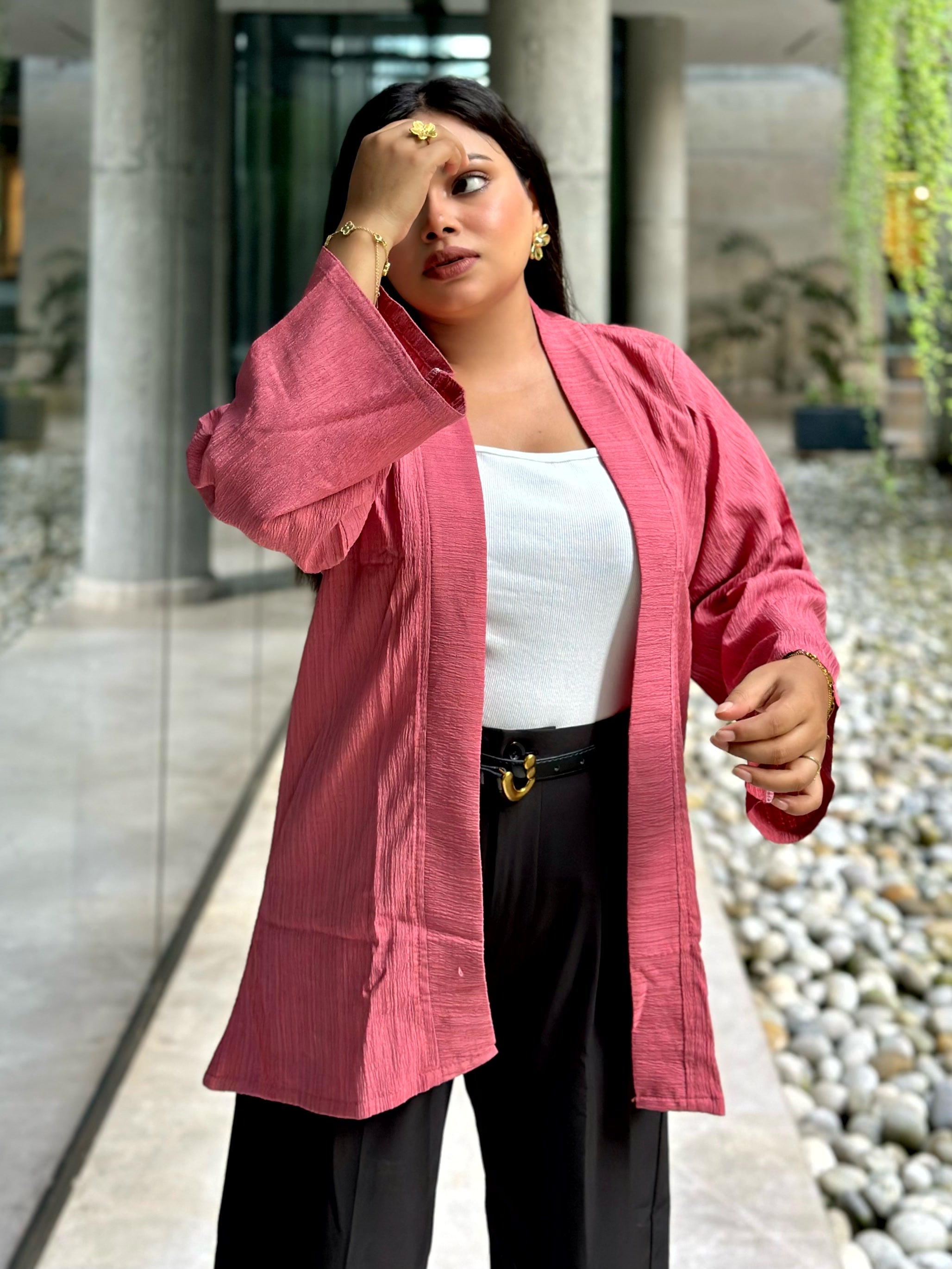 A woman wearing a pink textured open-front Kimono over a white top and black pants, standing in a modern outdoor walkway with stone columns and pebbles.