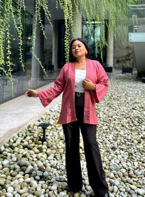 A woman stands outdoors on a bed of smooth river stones, wearing a pink open-front Kimono over a white top and black wide-leg pants. Hanging green vines drape over a modern glass building in the background.