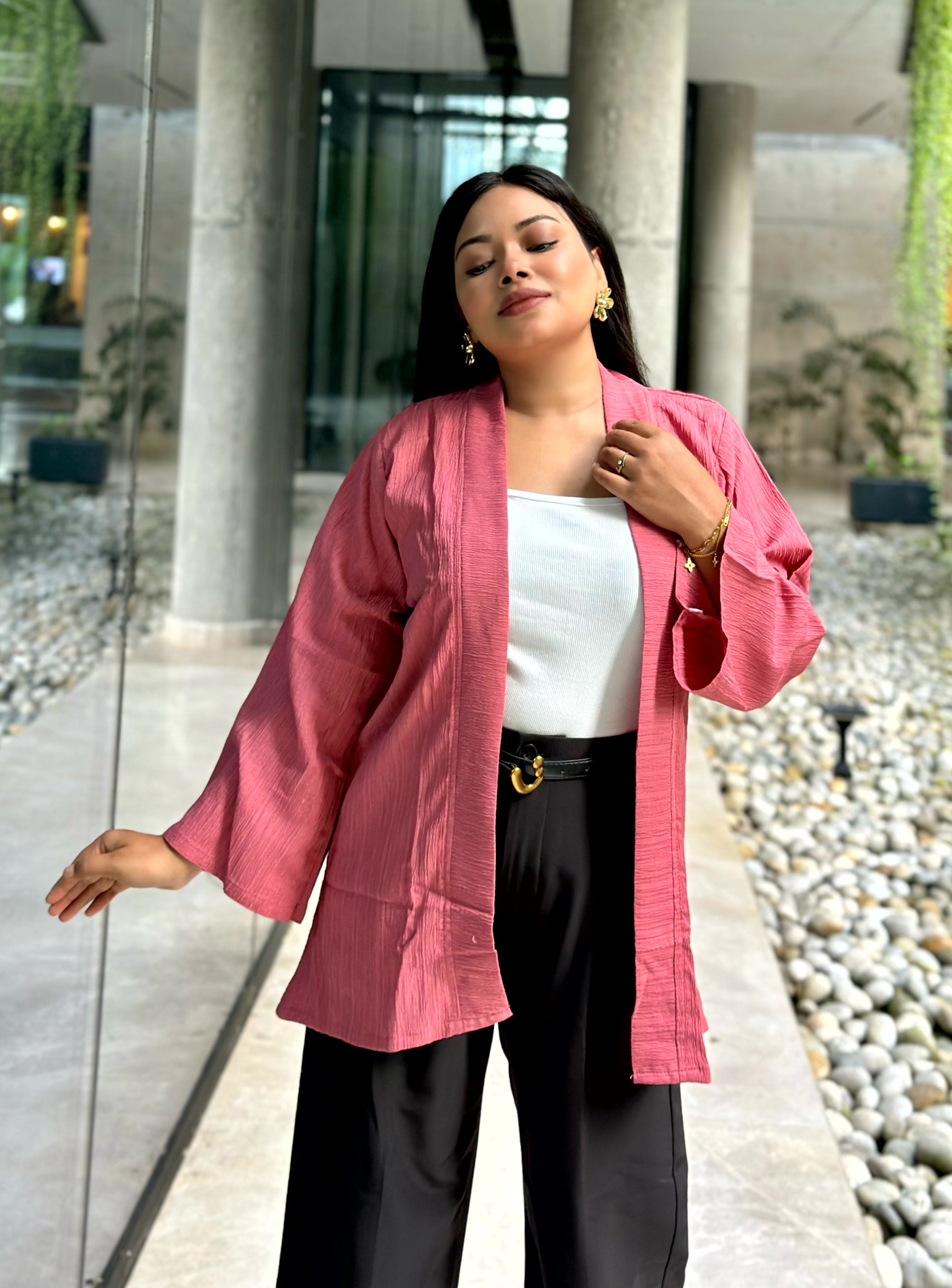 A woman wearing a pink textured open-front Kimono over a white top and black pants, standing in a modern outdoor walkway with stone columns and gravel.