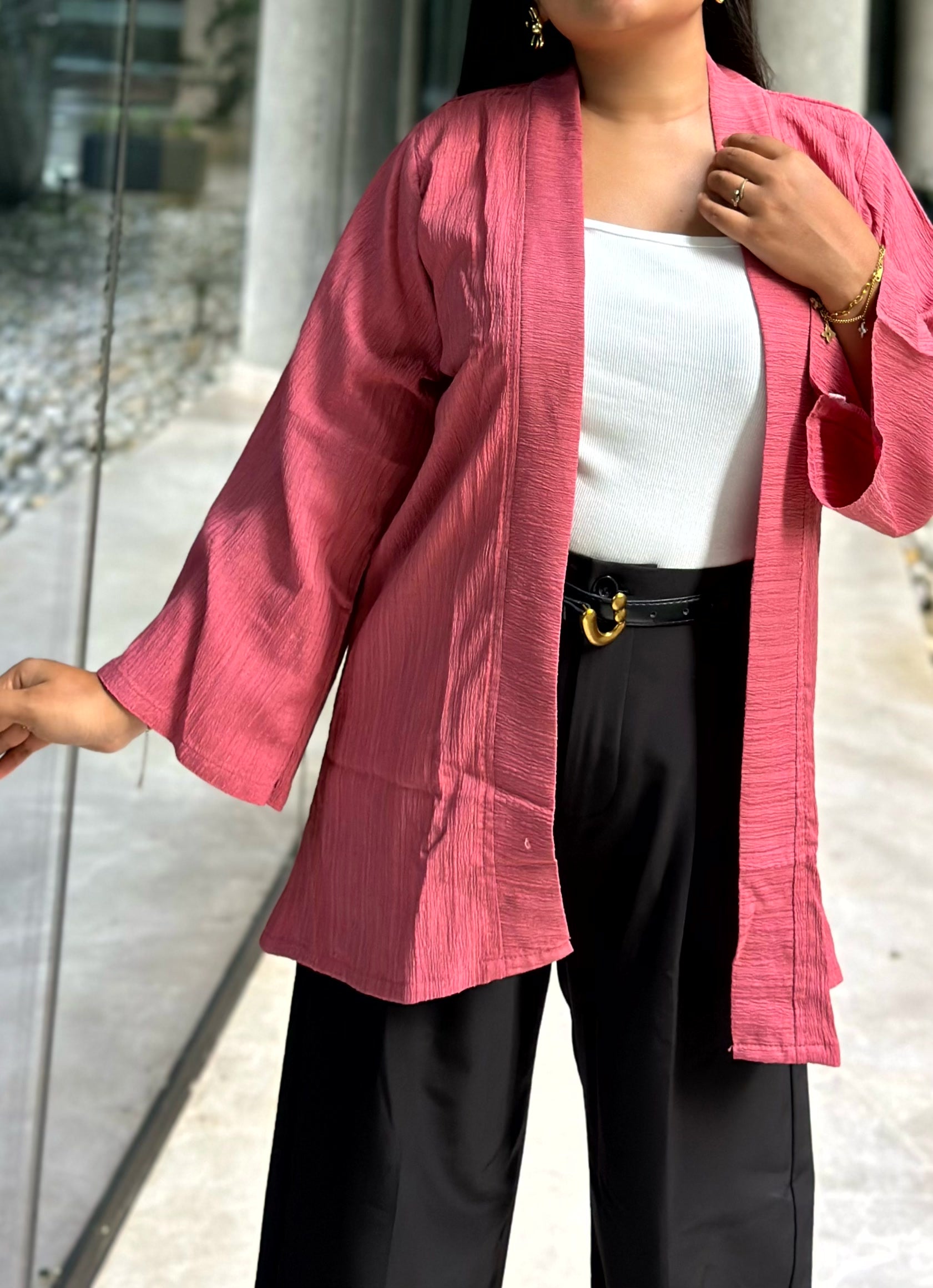 A woman wearing a pink textured open-front Kimono with wide sleeves over a white top and black pants, standing near a glass door.