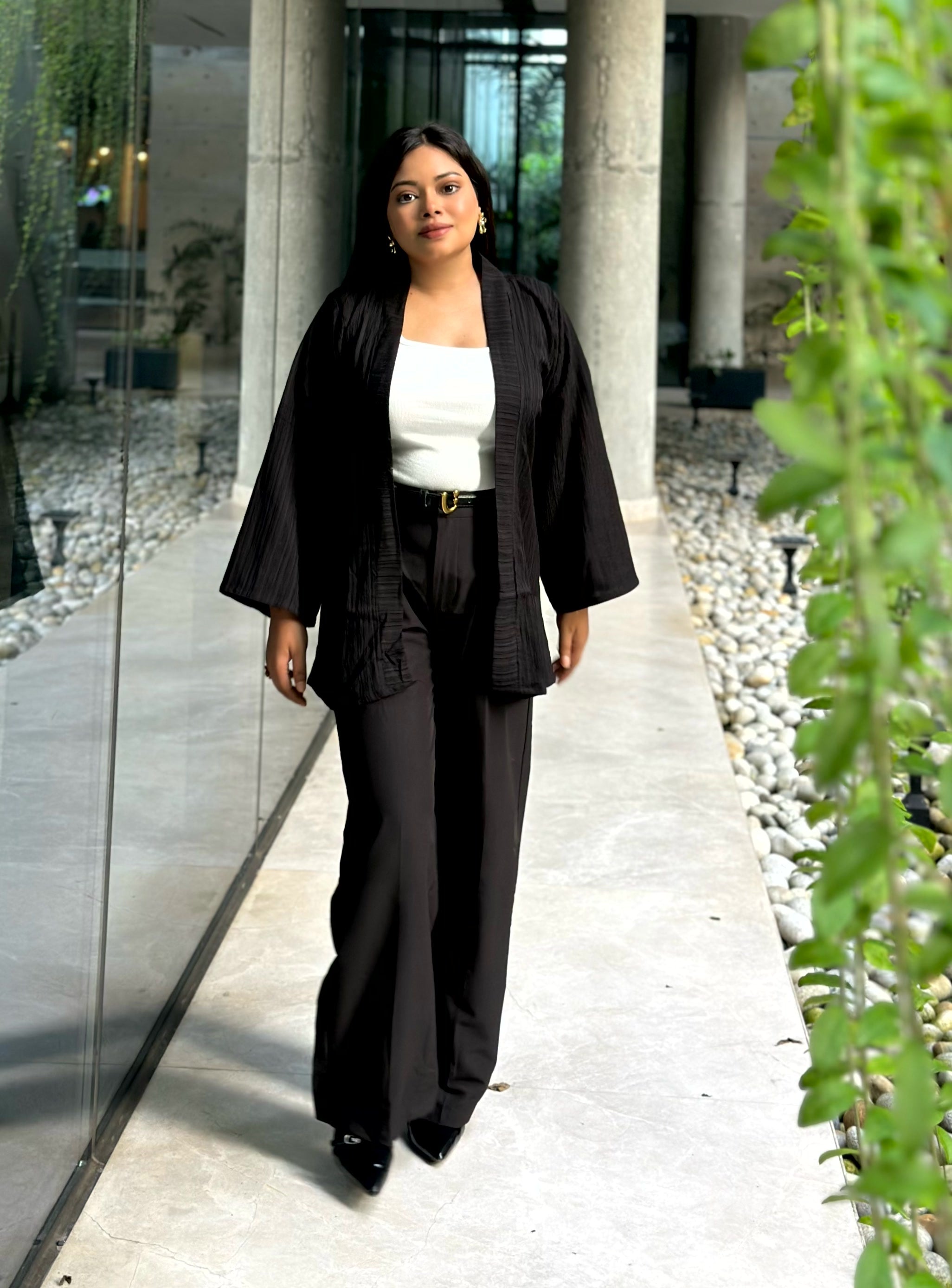 A woman walking down a modern outdoor walkway wearing a black textured kimono-style jacket, black wide-leg trousers, a white top, and black shoes.
