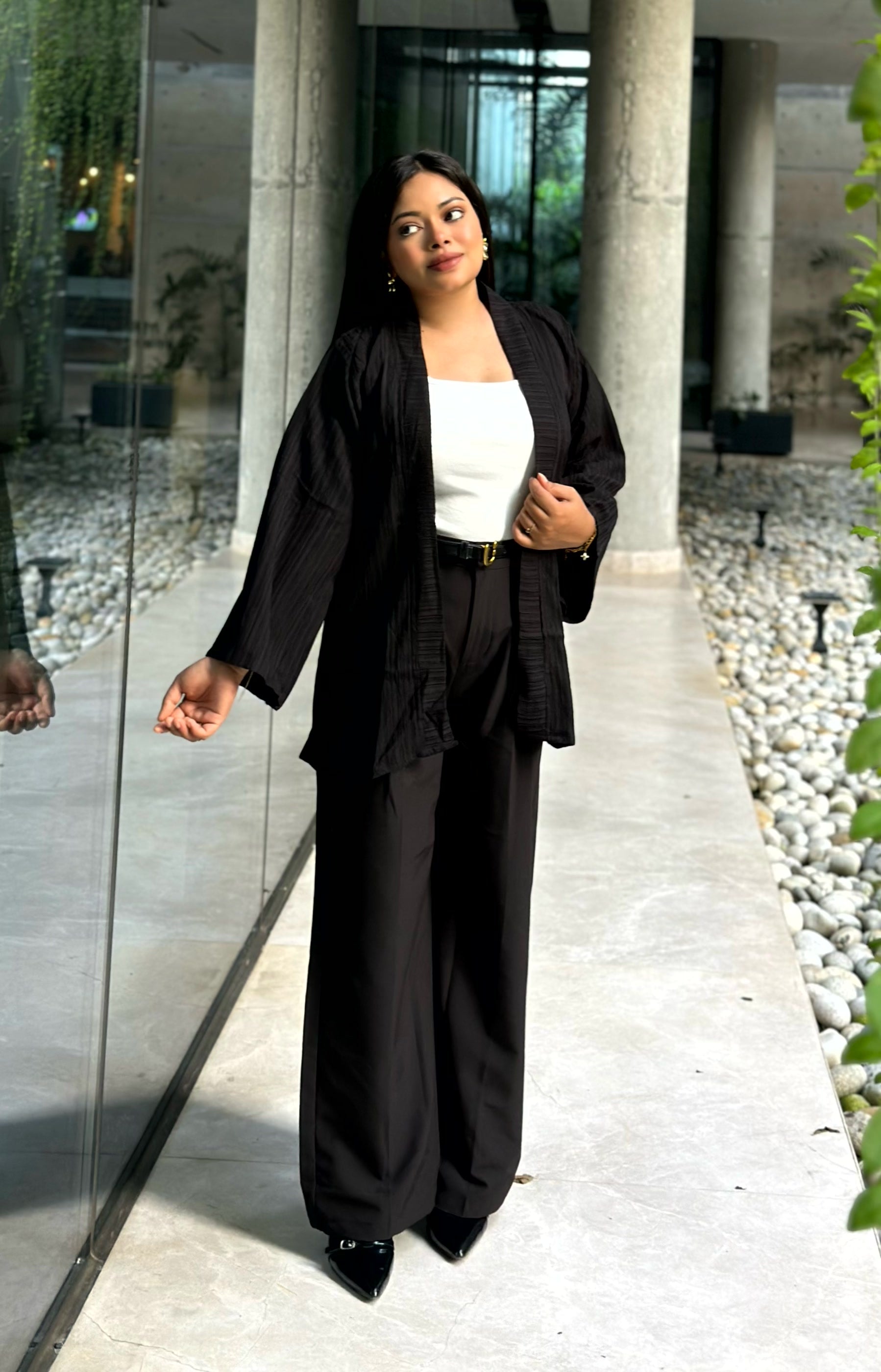A woman modeling a black two-piece outfit consisting of a long-sleeve open-front Kimono and matching wide-leg trousers, worn over a white top, in an outdoor walkway with stone columns and glass walls.