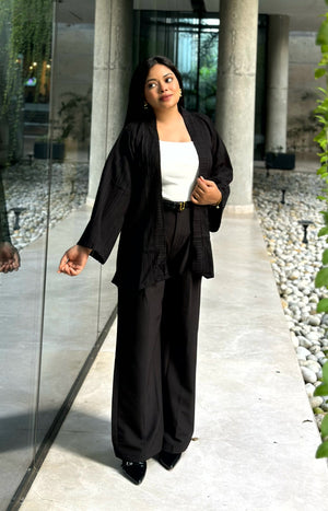 A woman modeling a black two-piece outfit consisting of a long-sleeve open-front Kimono and matching wide-leg trousers, worn over a white top, in an outdoor walkway with stone columns and glass walls.