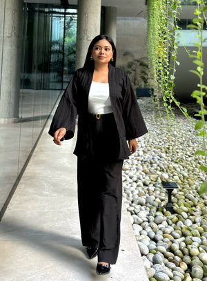 A woman walking outdoors in a modern architectural setting, wearing a black pinstriped Kimono and matching wide-leg trousers over a white top.