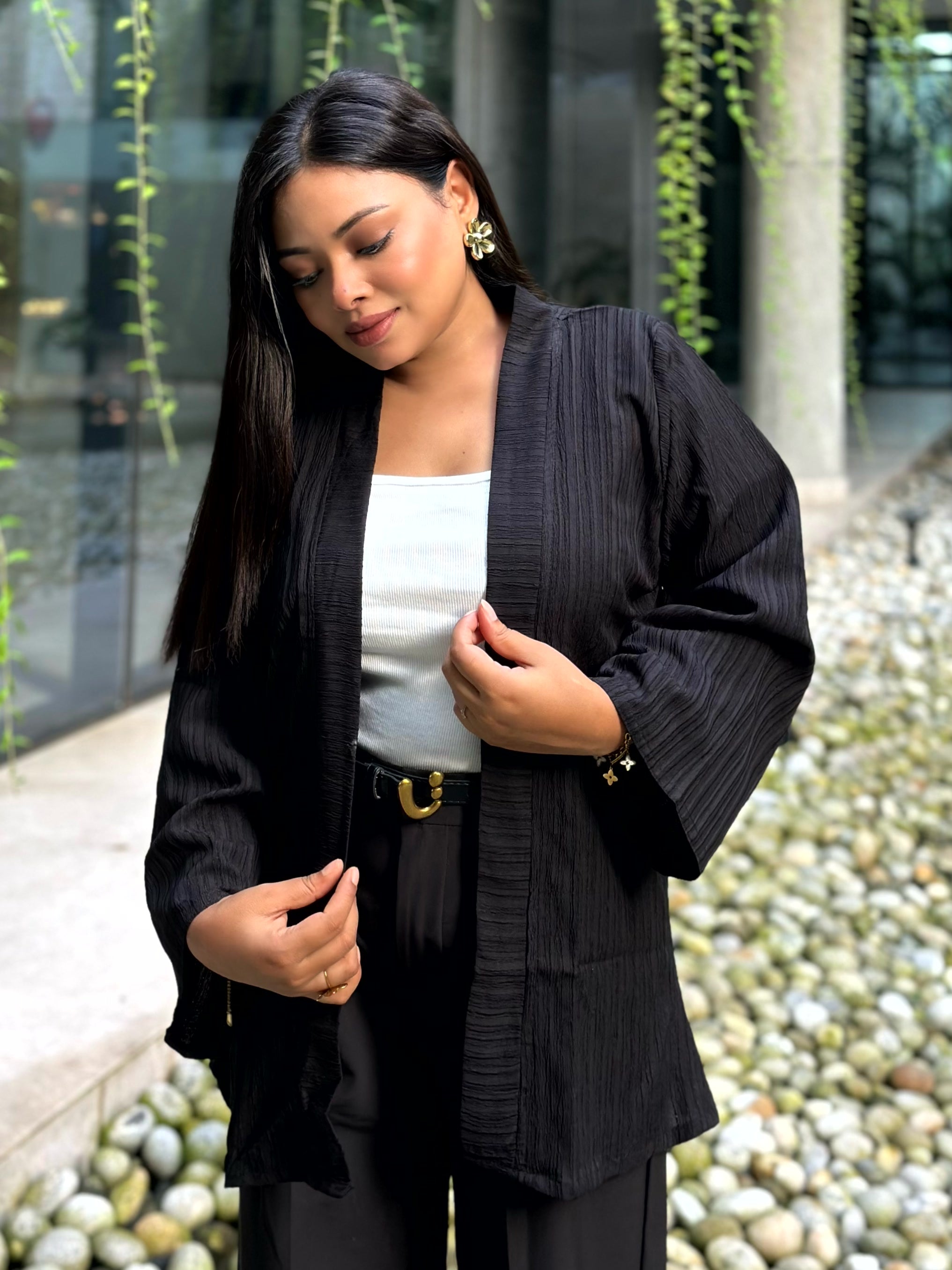 A woman wearing a black textured kimono-style jacket over a white top and black pants, standing outdoors near a glass building with greenery and pebbles in the background.