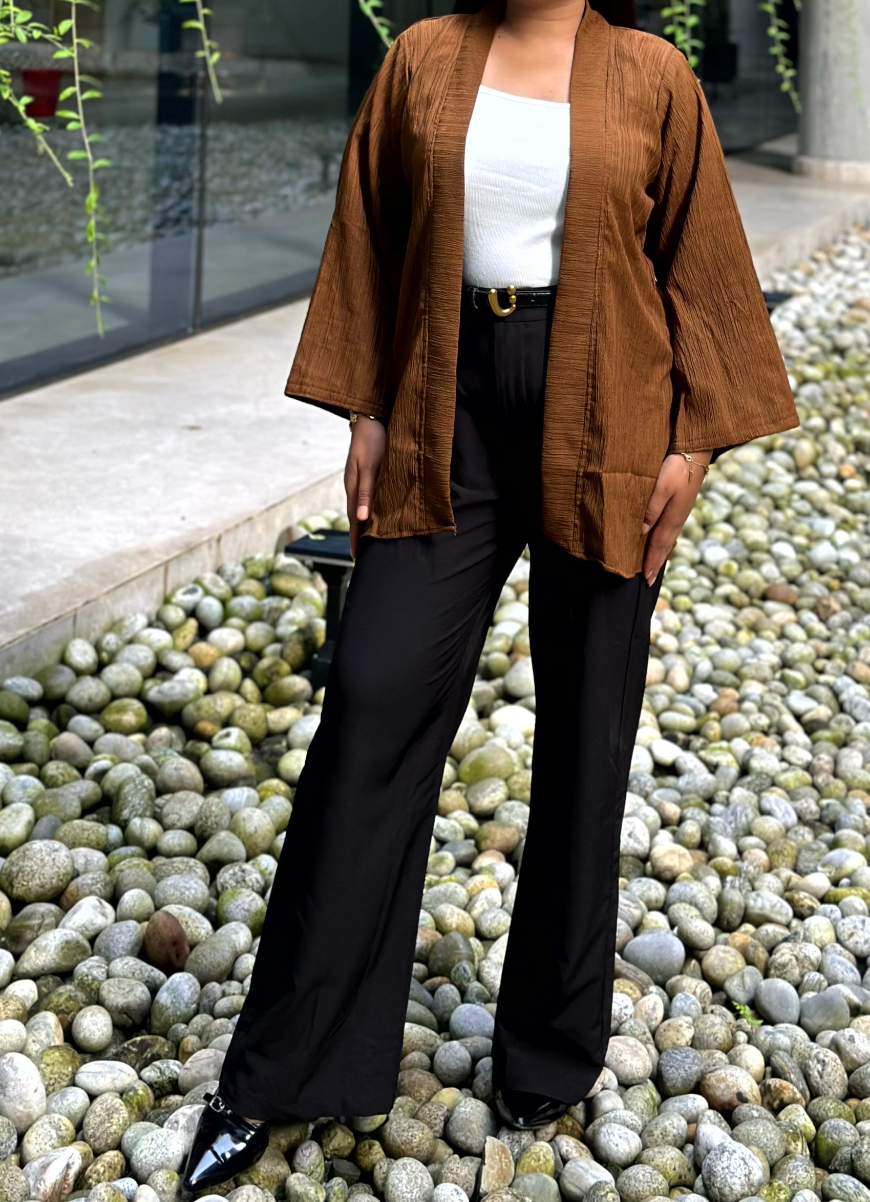 A woman wearing a brown open-front kimono with wide three-quarter sleeves, black wide-leg pants, and a white top, standing outdoors on a bed of smooth gray pebbles.