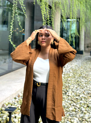 A woman wearing a brown textured open-front kimono over a white top and black pants, posing outdoors with her hands in front of her eyes. The background features a modern glass building, hanging green vines, and a stone pebble pathway.