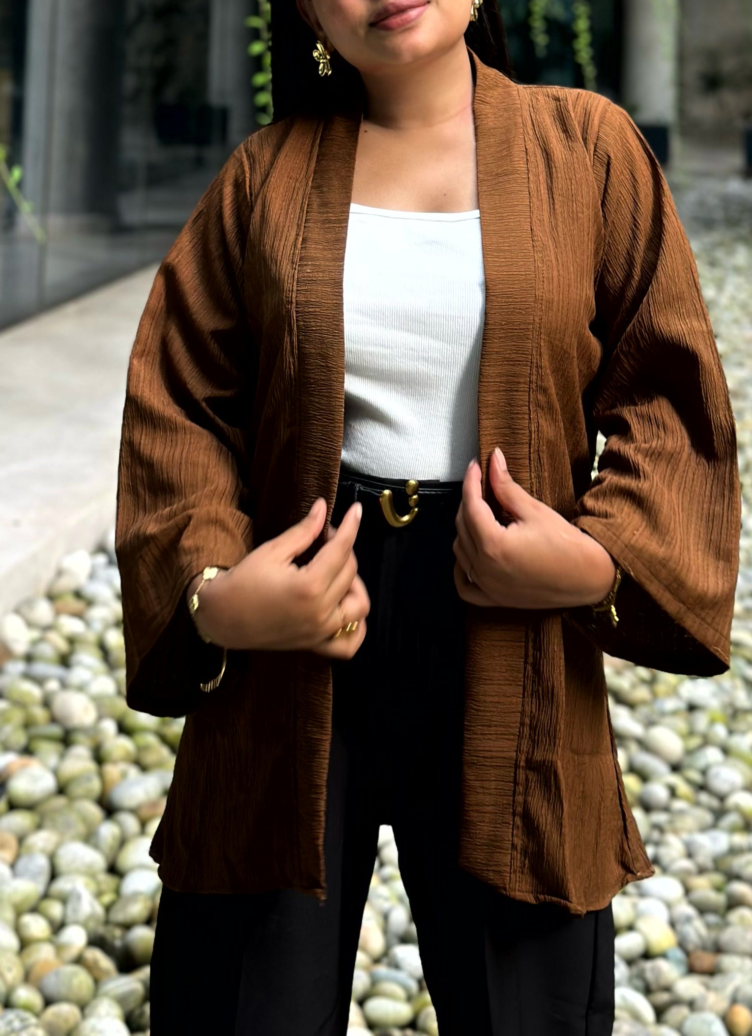A woman wearing a brown textured open-front kimono-style jacket over a white top and black pants, standing outdoors on a stone path.