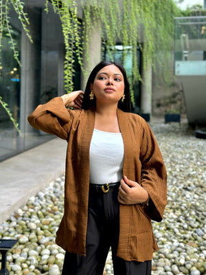 A woman wearing a brown textured open-front kimono over a white top and black pants, standing outdoors in a garden courtyard with hanging greenery and a stone pebble path.