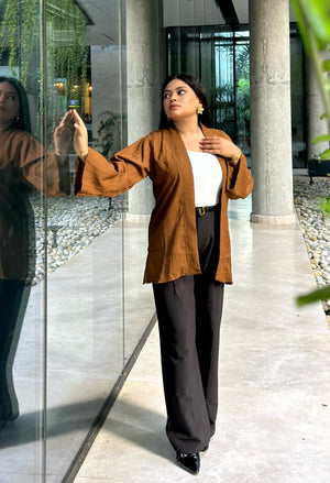 A woman models a brown open-front linen kimono with wide sleeves, worn over a white top and dark wide-leg trousers, standing in a modern indoor corridor with glass walls and columns.