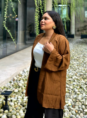 A woman wearing a brown textured open-front kimono over a white top and black pants, standing in an outdoor walkway with pebbles and green trailing plants.