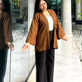 A woman modeling a brown open-front kimono with wide sleeves over a white top and black wide-leg trousers, standing in a modern walkway with glass walls and columns.