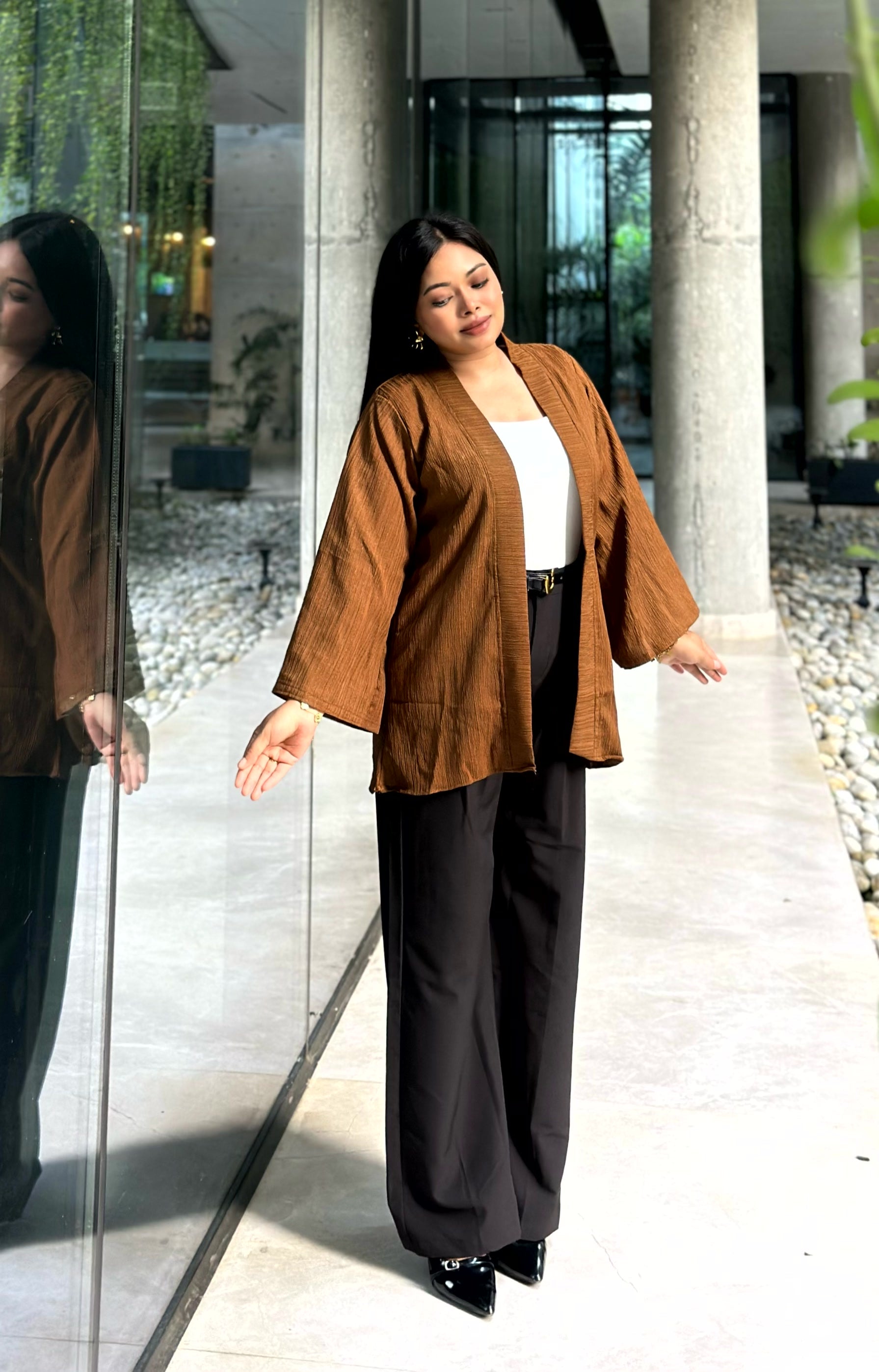 A woman modeling a brown open-front kimono with wide sleeves over a white top and black wide-leg trousers, standing in a modern walkway with glass walls and columns.