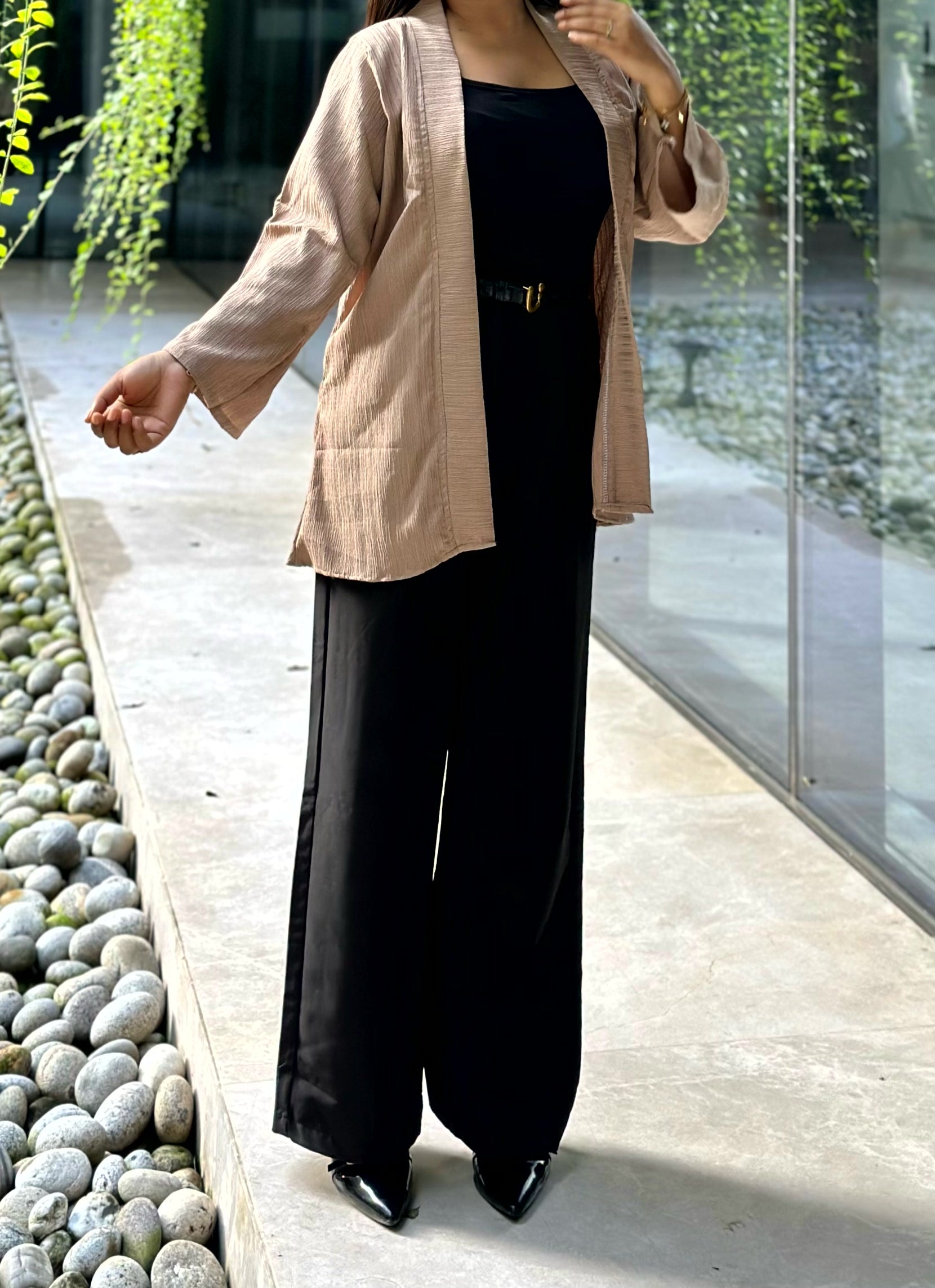 A woman wearing a beige textured open-front Kimono over a black top and black wide-leg pants, standing outdoors on a stone walkway next to a glass wall.