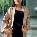 A woman wearing a light beige textured kimono-style Kimono over a black top and black skirt, standing in front of a glass wall with greenery visible outside.
