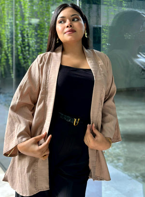 A woman wearing a light beige textured kimono-style Kimono over a black top and black skirt, standing in front of a glass wall with greenery visible outside.
