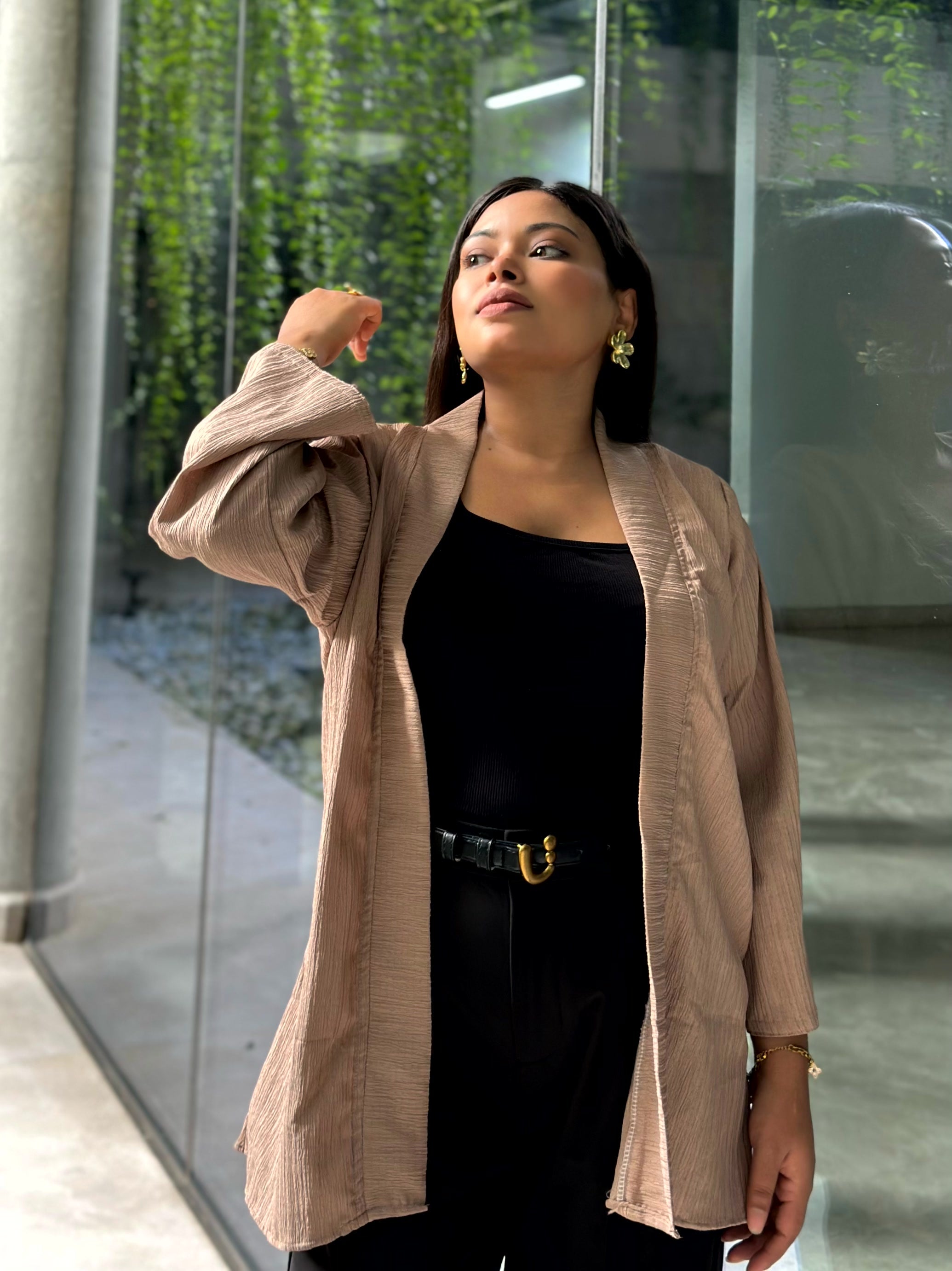 A woman wearing a light beige textured open-front Kimono over a black top and black pants with a gold-buckled belt, standing in front of a glass wall with greenery.