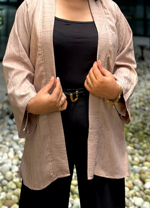 A person wearing a light beige textured open-front Kimono over a black top and black pants, standing outdoors in front of a stone wall.