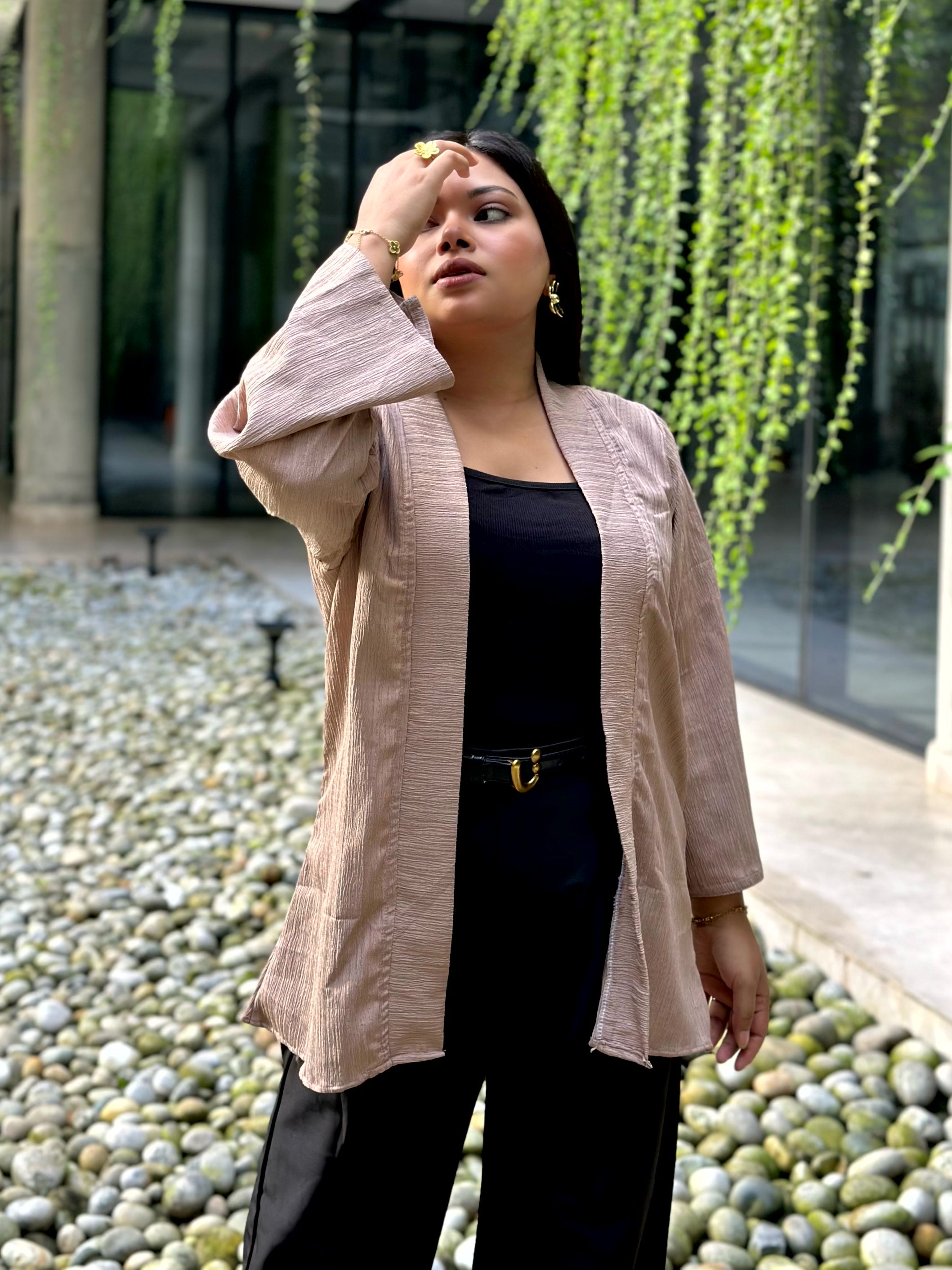 A woman wearing a light beige linen-textured open-front Kimono over a black top and black pants, standing outdoors in front of a modern building with green trailing plants.