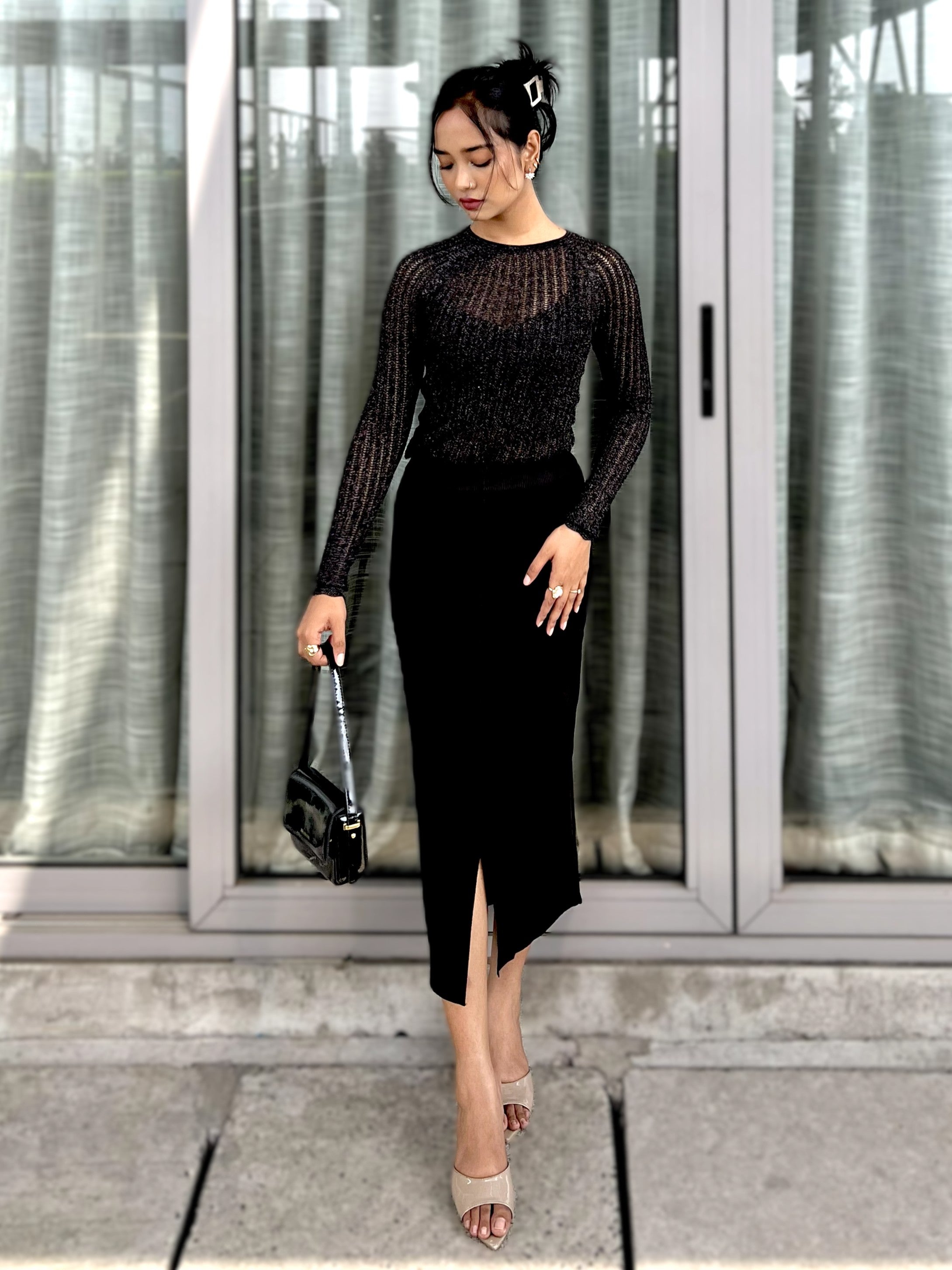 Woman in a black outfit with a mesh top and pencil skirt, holding a black handbag, standing in front of glass doors.