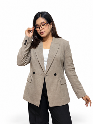 A woman modeling a light beige double-breasted blazer with black buttons, worn over a white top and black pants, against a plain white background.