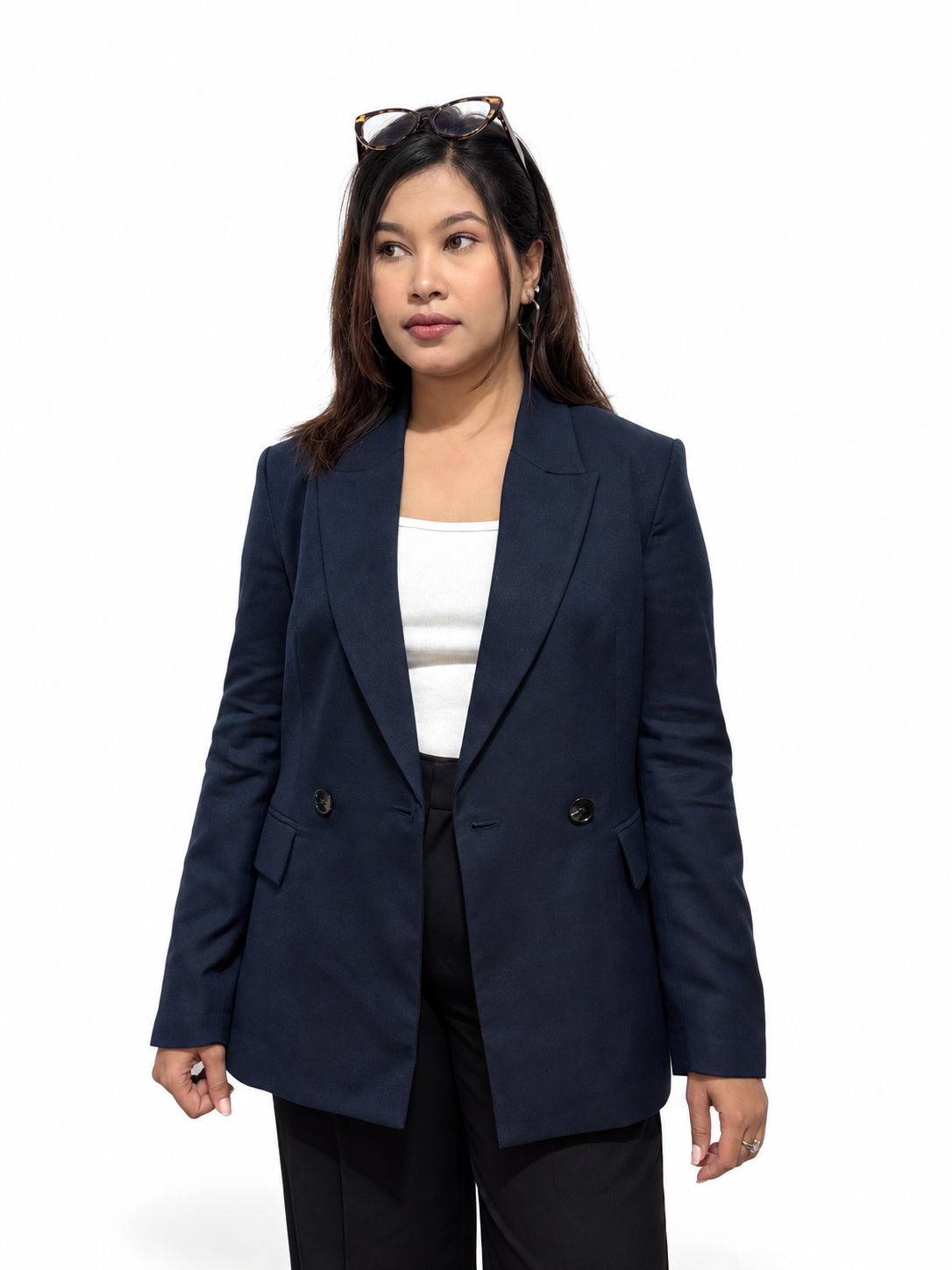 A woman modeling a navy blue double-breasted blazer over a white top and black trousers, standing against a plain white studio background with sunglasses on her head.
