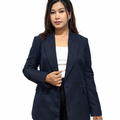 A woman modeling a navy blue double-breasted blazer over a white top and black pants, standing against a plain white studio background with sunglasses on her head and a small handbag in one hand.
