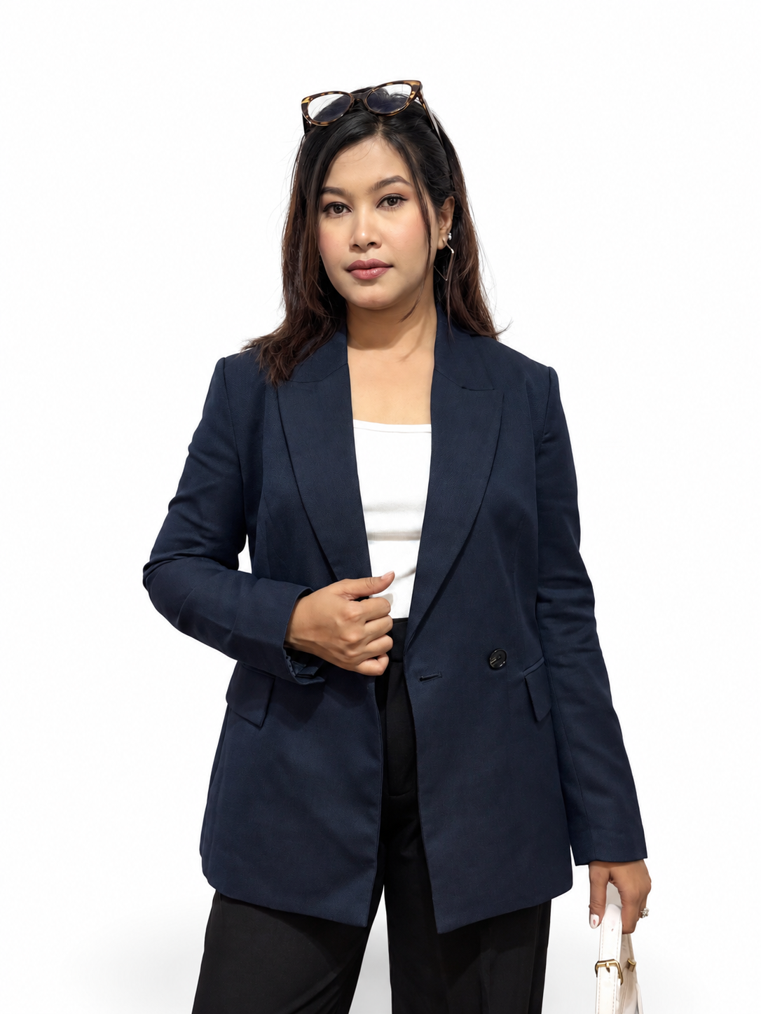 A woman modeling a navy blue double-breasted blazer over a white top and black pants, standing against a plain white studio background with sunglasses on her head and a small handbag in one hand.