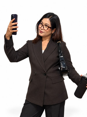 A woman wearing a dark brown double-breasted blazer and black pants takes a mirror selfie while holding a black shoulder bag and a black tumbler against a plain white background.