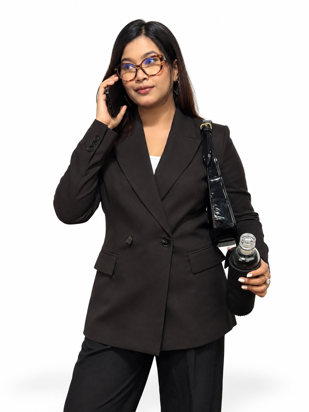 A woman wearing a tailored black double-breasted blazer and matching black trousers, holding a black shoulder bag and a bottle while talking on a phone against a plain white background.