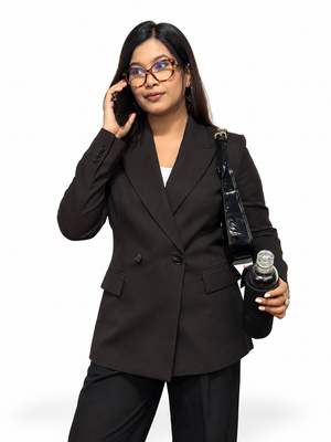 A woman wearing a tailored black double-breasted blazer and matching black trousers, holding a black shoulder bag and a bottle while talking on a phone against a plain white background.