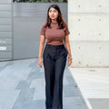 A woman walking outdoors on a paved walkway, wearing a fitted short-sleeve brown top and high-waisted black wide-leg trousers.