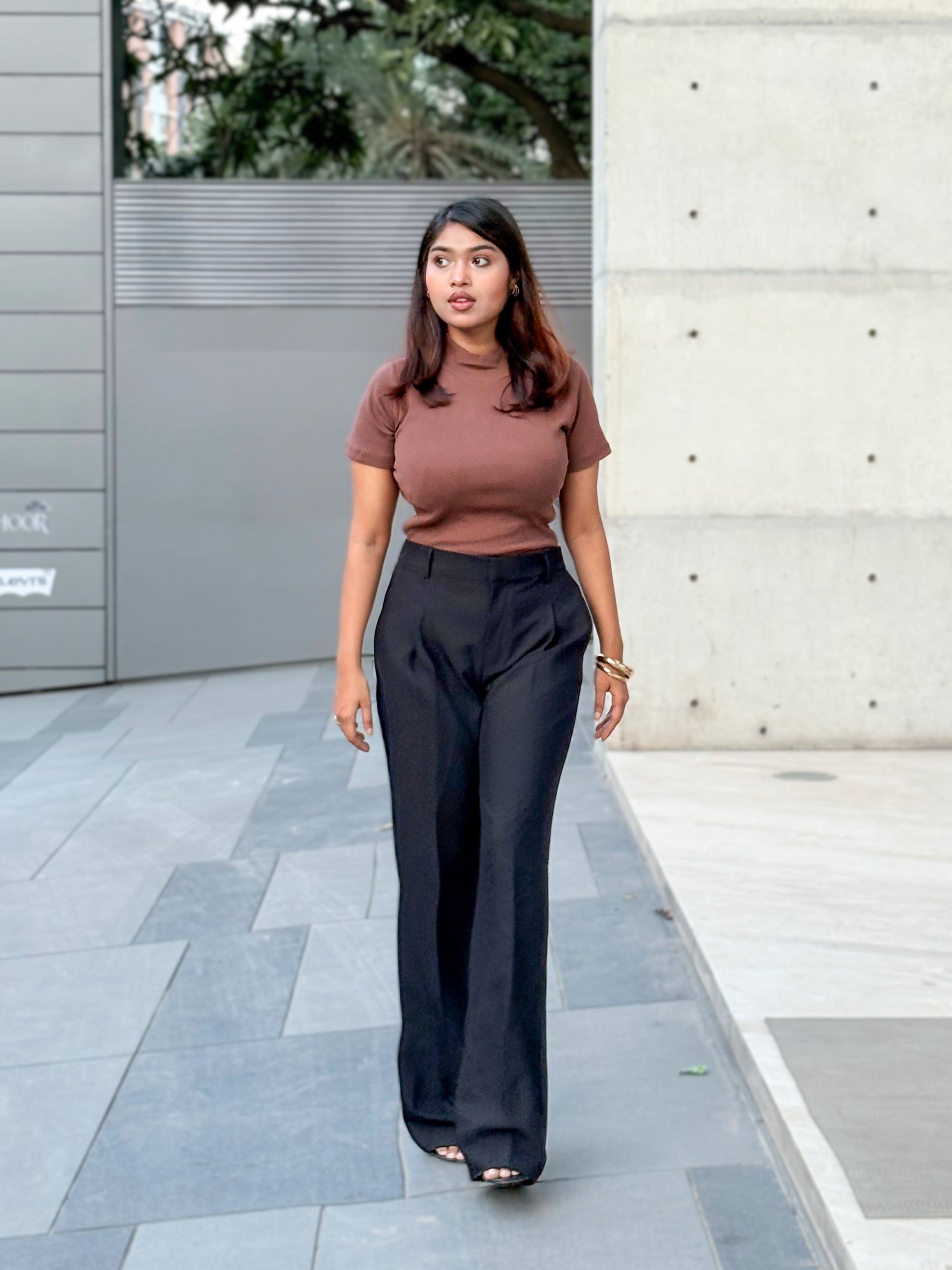 A woman walking outdoors on a paved walkway, wearing a fitted short-sleeve brown top and high-waisted black wide-leg trousers.