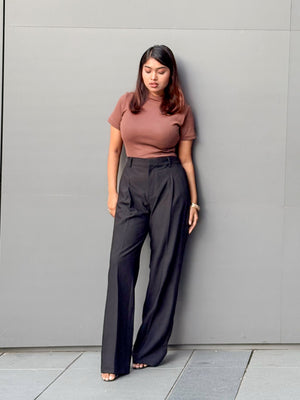 A woman stands against a gray wall wearing a fitted short-sleeve brown top and high-waisted black wide-leg trousers.