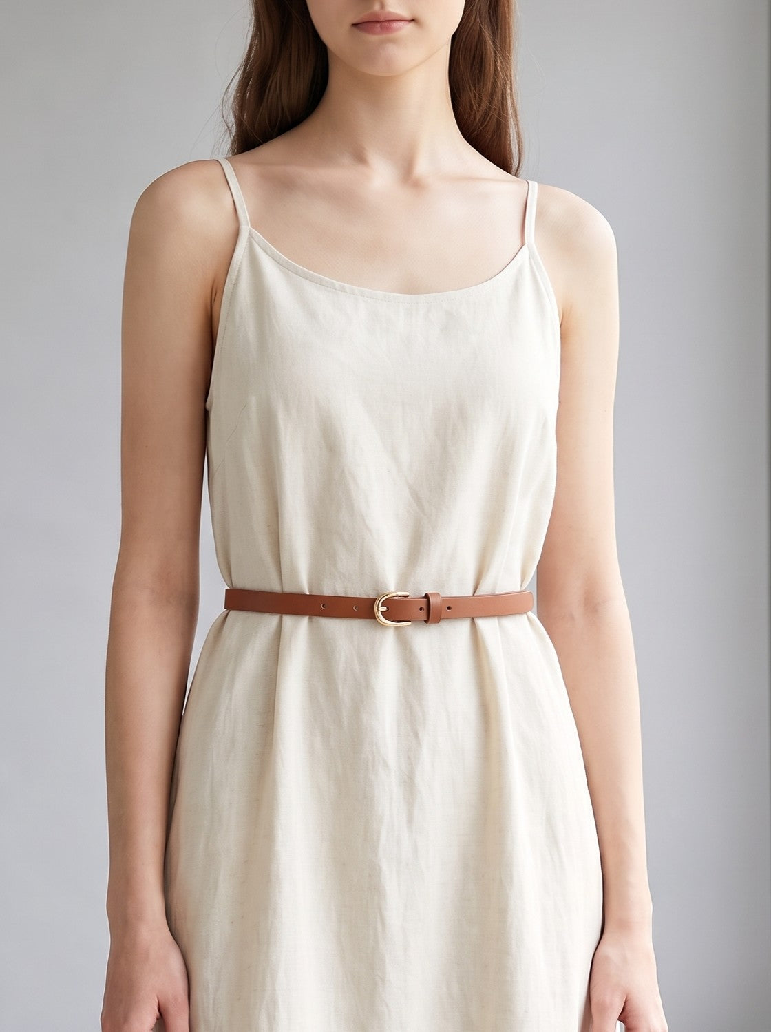 A woman wearing a cream-colored linen slip dress with thin spaghetti straps and a brown leather belt with a gold buckle, shown from the neck down against a neutral gray background.