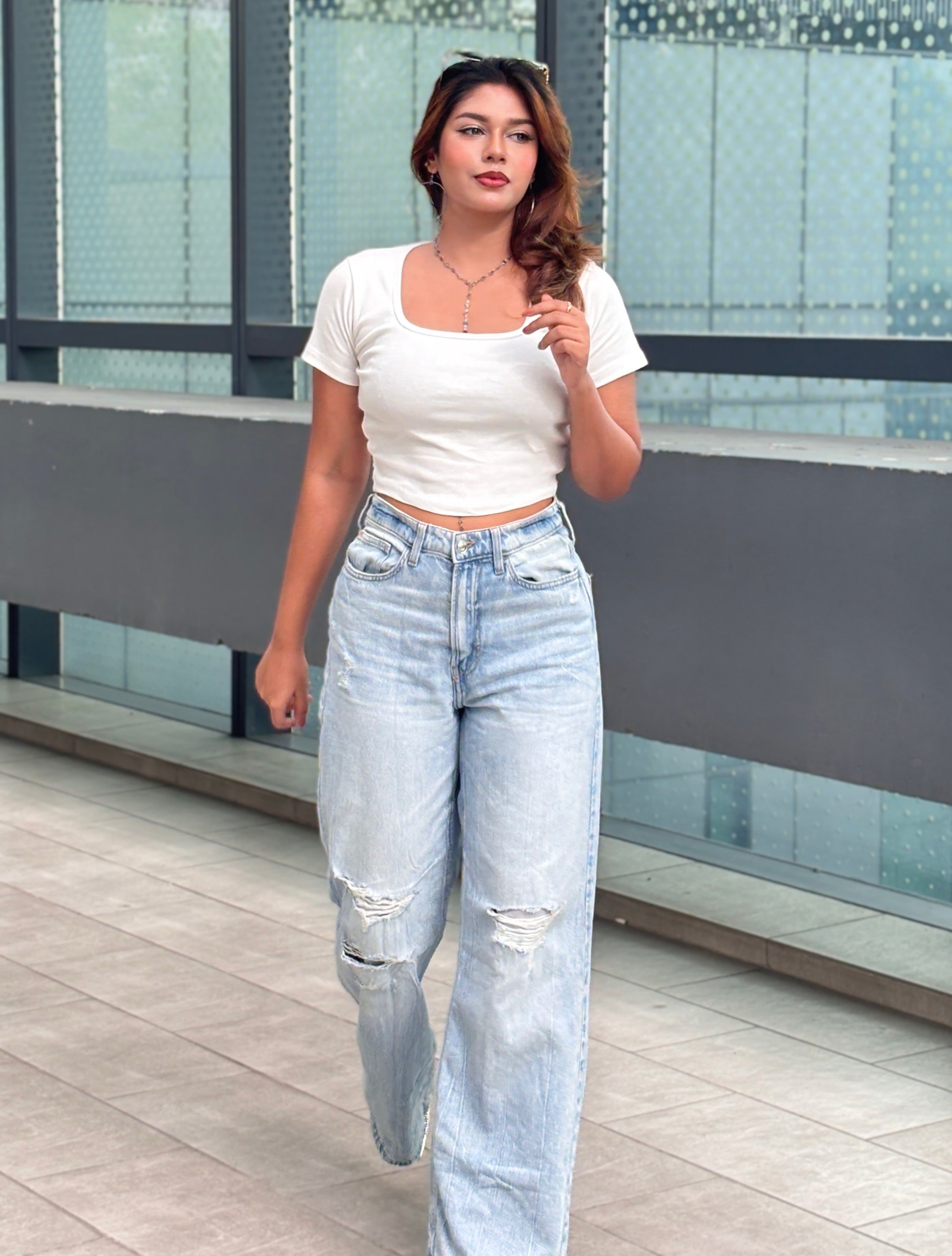 A woman walking outdoors wearing a white short-sleeve crop top and light-wash high-waisted wide-leg jeans with distressed details. TREND BUZZ