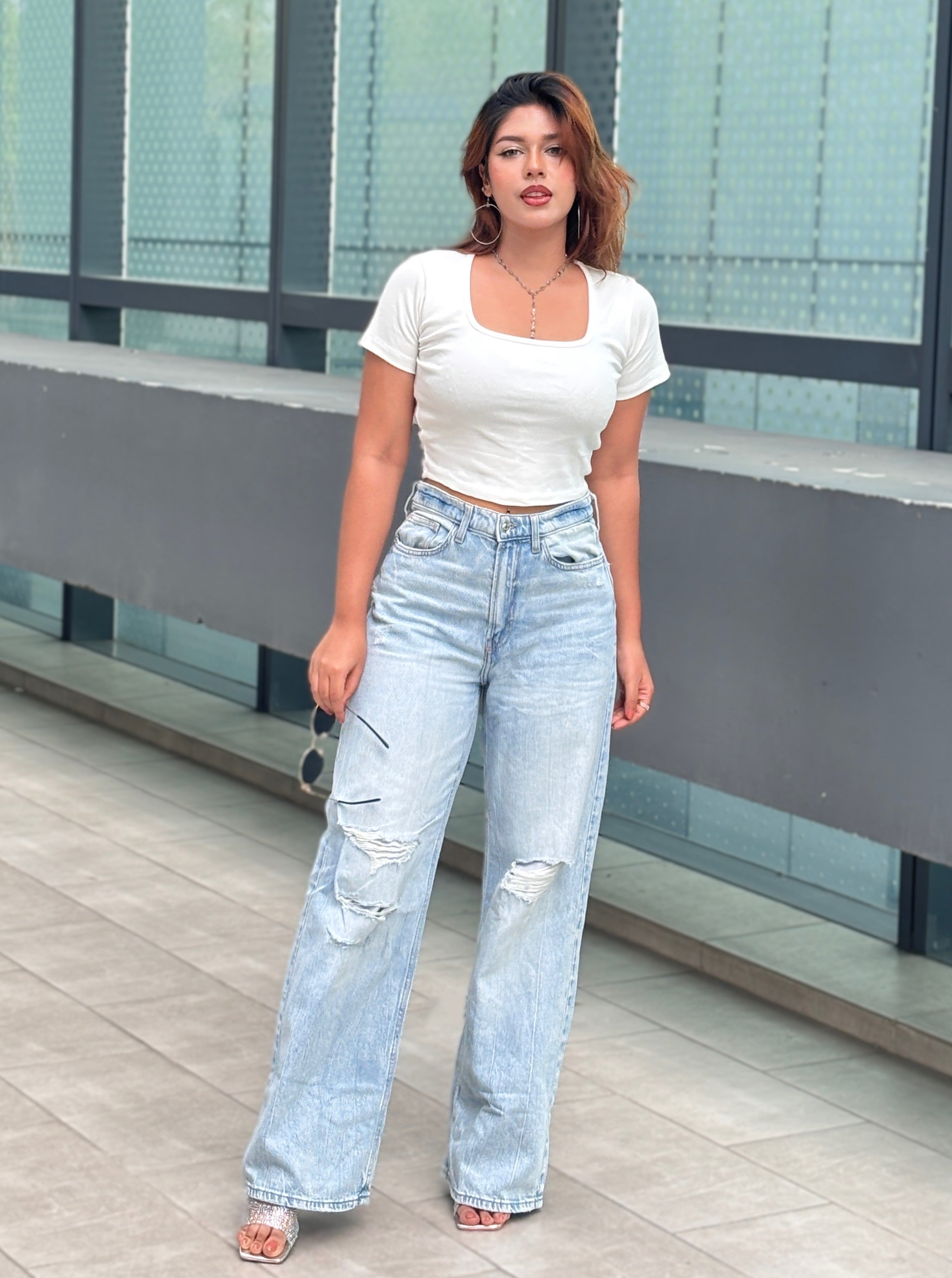 A woman modeling a white short-sleeve crop top and light-wash distressed wide-leg jeans, standing outdoors in front of a modern glass building.